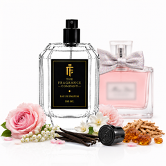 A clear faceted perfume bottle with a black spray top and black front label reading 'THE FRAGRANCE COMPANY' and 'EAU DE PARFUM 100 ML'. A pink perfume bottle with a silver bow sits behind it.