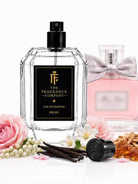 A clear faceted perfume bottle with a black spray top and black front label reading 'THE FRAGRANCE COMPANY' and 'EAU DE PARFUM 100 ML'. A pink perfume bottle with a silver bow sits behind it.