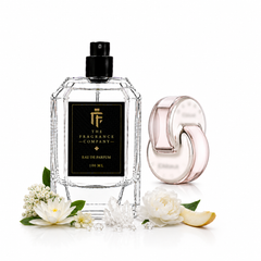 A clear glass perfume bottle with a black cap and black label reading 'The Fragrance Company Eau de Parfum 100 ML' stands beside a pale pink translucent spray atomizer. White flowers, green leaves, and a small slice of citrus are arranged around them on a clean white background.