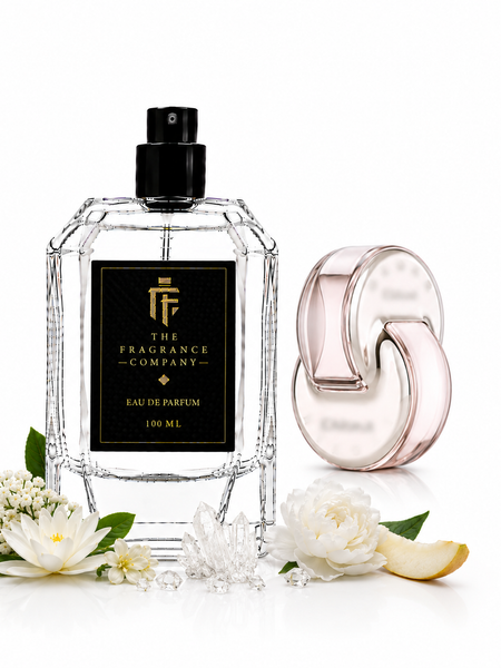 A clear glass perfume bottle with a black cap and black label reading 'The Fragrance Company Eau de Parfum 100 ML' stands beside a pale pink translucent spray atomizer. White flowers, green leaves, and a small slice of citrus are arranged around them on a clean white background.