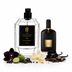 Two perfume bottles from The Fragrance Company are displayed on a white background with botanical and food props. The main bottle is a faceted clear glass bottle with a black cap and a black label featuring a gold logo and text.