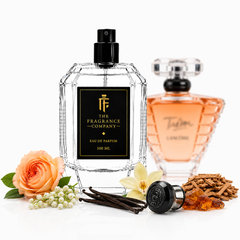 A clear faceted perfume bottle with a black spray top and black label reading 'THE FRAGRANCE COMPANY' stands on a white studio background. A second amber-toned perfume bottle is blurred in the back right, and decorative fragrance-themed props including a peach rose, white flower, vanilla pods, cinnamon sticks, and amber pieces are arranged around the bottles.