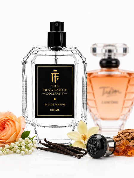 A clear faceted perfume bottle with a black spray top and black label reading 'THE FRAGRANCE COMPANY' stands on a white studio background. A second amber-toned perfume bottle is blurred in the back right, and decorative fragrance-themed props including a peach rose, white flower, vanilla pods, cinnamon sticks, and amber pieces are arranged around the bottles.