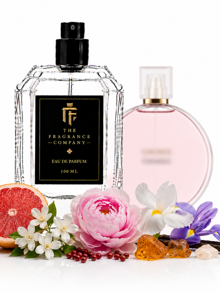 A clear glass perfume bottle with a black spray top and a black label reading 'THE FRAGRANCE COMPANY' stands in front of a pale pink perfume bottle. The bottle is styled with a sliced grapefruit, a pink flower, white blossoms, purple flowers, amber crystals, and vanilla beans on a white background.