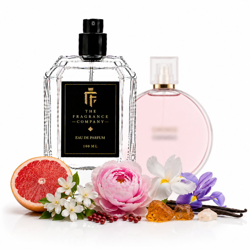 A clear glass perfume bottle with a black spray top and a black label reading 'THE FRAGRANCE COMPANY' stands in front of a pale pink perfume bottle. The bottle is styled with a sliced grapefruit, a pink flower, white blossoms, purple flowers, amber crystals, and vanilla beans on a white background.