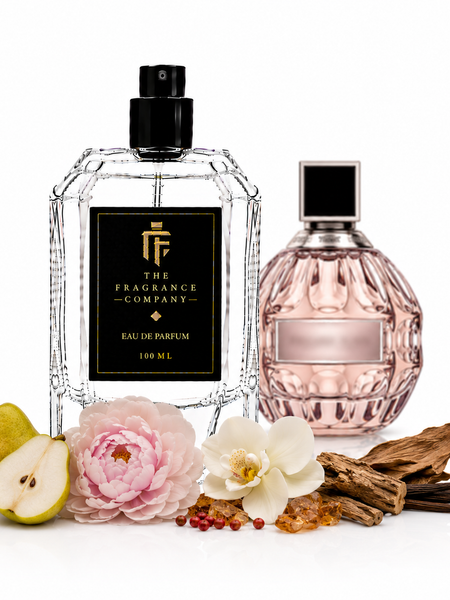 Two perfume bottles from The Fragrance Company on a white background, styled with pear halves, pink and white flowers, vanilla pods, and amber beads. The left bottle is clear faceted glass with a black spray top and black label.