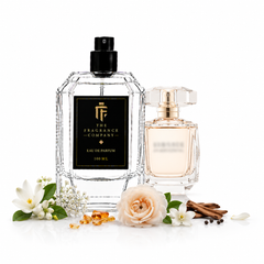 Two perfume bottles from The Fragrance Company on a white background, styled with white flowers, cinnamon sticks, and amber droplets. The larger bottle on the left is clear glass with a black label and black cap, and the smaller bottle on the right has a pale pink liquid and a gold cap.