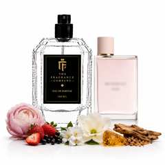 A clear glass perfume bottle with a black label reading 'The Fragrance Company' stands in front of a pale pink rectangular perfume bottle. The main bottle is styled with flowers, berries, wood, and seeds on a clean white background.