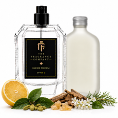 A clear glass perfume bottle with a black spray top and a black label reading 'THE FRAGRANCE COMPANY' stands on a white background, styled with a sliced orange, green leaves, bark, white flowers, and a frosted bottle behind it.