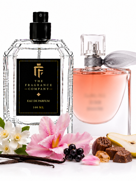 Two perfume bottles from The Fragrance Company on a white background, styled with floral and fruit props including white and pink flowers, vanilla beans, blackberries, and pear slices. The left bottle is clear glass with a black label and black spray top, and the right bottle is a soft pink glass spray bottle with a gray ribbon.