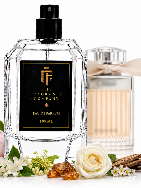 A 100 ml clear glass perfume bottle with a black spray top and a black label featuring gold lettering, displayed alongside a second ribbed glass perfume bottle in the background. The main bottle is styled with a pink rose, white rose, vanilla beans, amber crystals, and small white flowers on a white background.
