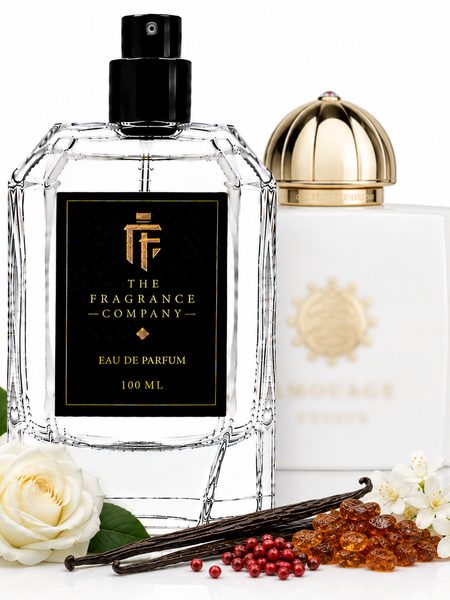Two perfume bottles from The Fragrance Company on a white background, styled with white roses, vanilla beans, and amber beads. The foreground bottle is a faceted clear glass bottle with a black cap and a black label featuring gold lettering.