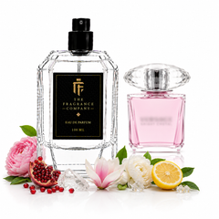 Two perfume bottles from The Fragrance Company on a white background, styled with flowers, a pomegranate, and a lemon. The larger bottle on the left is clear glass with a black spray top and a black label featuring gold lettering.