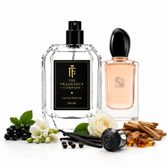 Two elegant perfume bottles on a white reflective surface with botanical and spice props. The front bottle is a faceted clear glass bottle with a black spray top and a black label reading 'THE FRAGRANCE COMPANY' and 'EAU DE PARFUM 100 ML'.