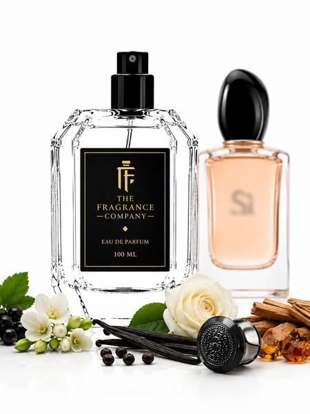 Two elegant perfume bottles on a white reflective surface with botanical and spice props. The front bottle is a faceted clear glass bottle with a black spray top and a black label reading 'THE FRAGRANCE COMPANY' and 'EAU DE PARFUM 100 ML'.