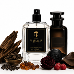 A clear glass perfume bottle labeled 'The Fragrance Company' with a black spray top and black label, styled with dark amber liquid in a bottle behind it, plus rose petals, dried bark, peppercorns, amber beads, and raspberries on a white background.