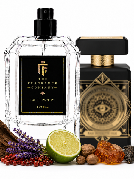 A clear faceted perfume bottle labeled 'The Fragrance Company' stands in front of a black-and-gold branded box, with botanical and spice ingredients arranged around the base on a white background.