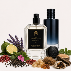 A clear glass perfume bottle with a black label reading 'THE FRAGRANCE COMPANY' stands centered on a white surface, surrounded by fragrance ingredients including lavender, lime, peppercorns, berries, roots, bark, and an oyster shell. A dark blue bottle and a round black cap are visible behind it against a clean white background.